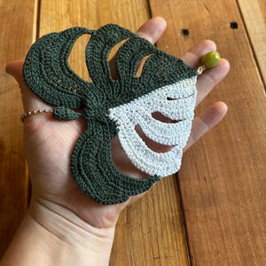 May include: A crocheted green and white monstera leaf with a green bead attached to the top.