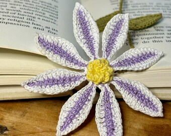 Op de afbeelding: Een gehaakt bladwijzer in de vorm van een bloem met witte bloemblaadjes en paarse accenten. Het midden van de bloem is geel.