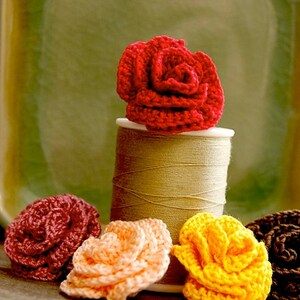 Puede incluir: Cinco flores de ganchillo en diferentes colores, incluyendo rojo, rosa, amarillo y marr&oacute;n, est&aacute;n dispuestas sobre una superficie de madera. Las flores est&aacute;n sentadas encima de un carrete de hilo.