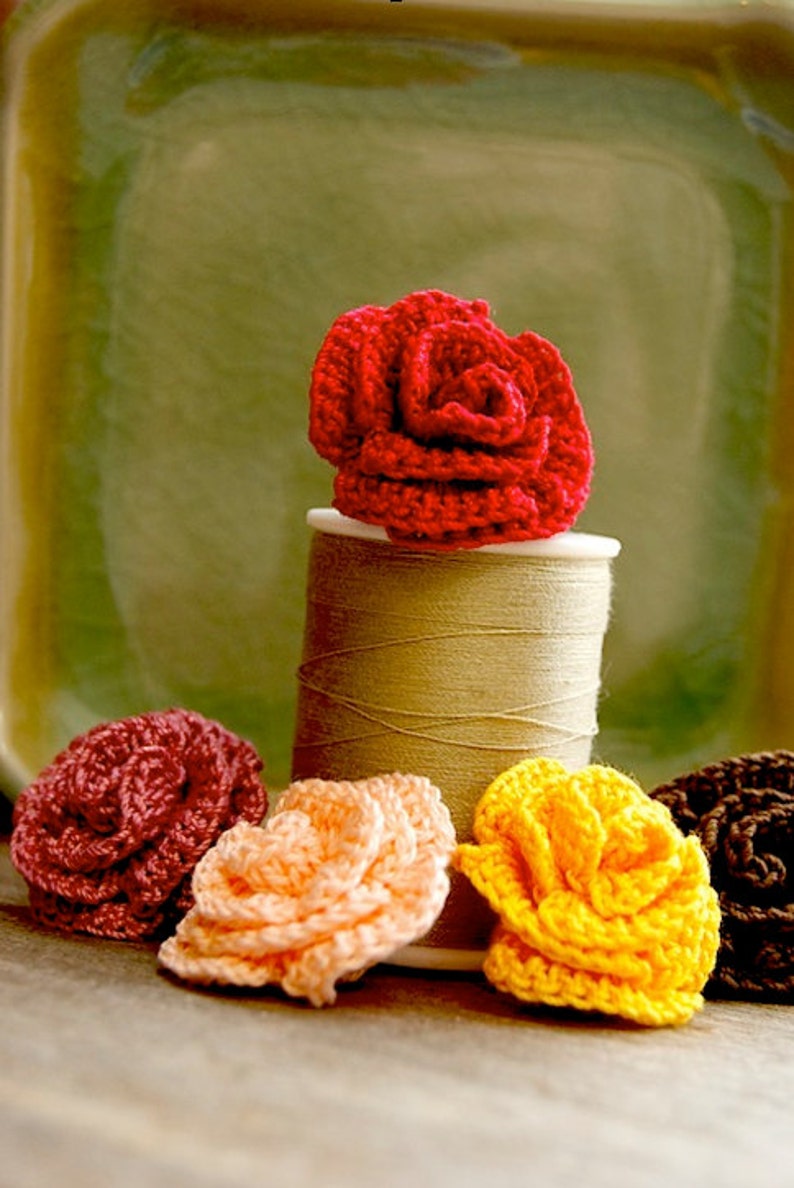 Puede incluir: Cinco flores de ganchillo en diferentes colores, incluyendo rojo, rosa, amarillo y marr&oacute;n, est&aacute;n dispuestas sobre una superficie de madera. Las flores est&aacute;n sentadas encima de un carrete de hilo.