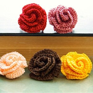 Puede incluir: Cuatro flores de rosa de ganchillo en diferentes colores: rojo, rosa, melocot&oacute;n y amarillo. Las flores est&aacute;n dispuestas sobre una superficie de madera y una bandeja de cer&aacute;mica.