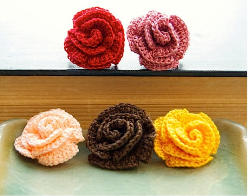 Puede incluir: Cuatro flores de rosa de ganchillo en diferentes colores: rojo, rosa, melocot&oacute;n y amarillo. Las flores est&aacute;n dispuestas sobre una superficie de madera y una bandeja de cer&aacute;mica.