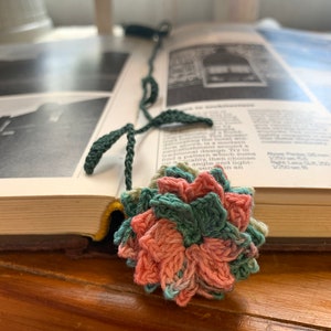 May include: A crocheted flower bookmark with pink, green, and blue yarn. The bookmark is attached to a green yarn stem with leaves.
