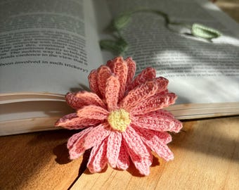 Op de afbeelding: Een gehaakte roze bloem met een gele kern, liggend op een houten oppervlak. De bloem is een bladwijzer en ligt op een open boek.