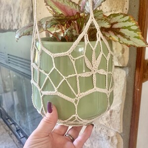 May include: A green ceramic planter with a white crocheted hanging planter. The plant has green and pink leaves.