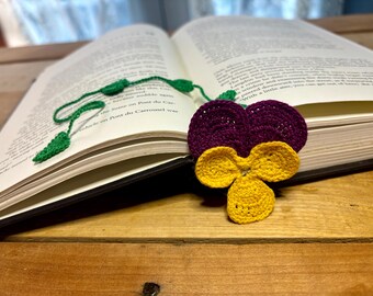 Op de afbeelding: Een gehaakt paars en geel viooltje boekenlegger met groene bladeren. De boekenlegger is geplaatst in een open boek.