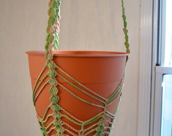 Op de afbeelding: Een terracotta plantenpot die hangt aan een gehäkelde macrame plantenhanger met groen en roze garen.