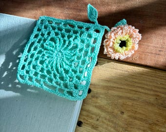 Puede incluir: Un marcapáginas cuadrado de ganchillo turquesa con un detalle de hoja y una flor de ganchillo melocotón y amarillo. Los artículos hechos a mano están sobre una superficie de madera junto a un libro.