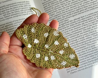 Op de afbeelding: Een gehaakt groen blad met witte stippen, vastgehouden in een hand. Het blad is een handgemaakt ambachtelijk item, perfect om een vleugje natuur aan uw interieur toe te voegen.