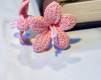 Op de afbeelding: Twee roze gehaakte bloemen, waarvan er één iets groter is dan de andere, liggen op een wit oppervlak. De bloemen zijn gemaakt met een delicate steek en hebben een zachte, vrouwelijke uitstraling.