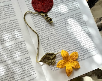 Op de afbeelding: Een gehaakte bladwijzer met een rode appel, groene bladeren en een gele bloem. De bladwijzer ligt op een open boek met de zichtbare tekst "The Wesleys Believe Their Is Haunted".