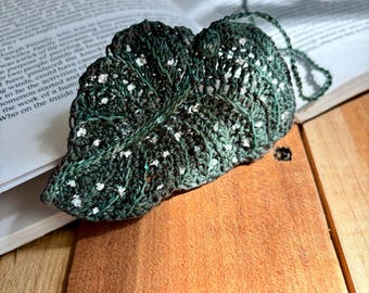 Op de afbeelding: Een groene gehaakte blad bladwijzer met witte accenten. De bladwijzer ligt op een houten oppervlak.