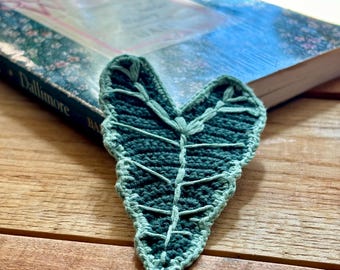 Op de afbeelding: Een handgemaakte, groene gehaakte bladwijzer in de vorm van een blad ligt op een boek met een blauwe kaft. De bladwijzer heeft een gedetailleerd ontwerp van bladnerven en een lichtgroene rand. Het boek is open, met de titel "Dallimore" zichtbaar.