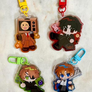 May include: Four acrylic keychains featuring chibi-style characters. One has a television head, one a dark-haired figure with a rabbit, one a green-eyed character with an eyepatch, and one a blue-eyed character holding a large blade.