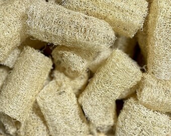 May include: A basket filled with several natural loofah sponges. The loofahs are cylindrical, beige, and have a textured, fibrous appearance. The basket is woven from light brown material, and the loofahs are densely packed within.