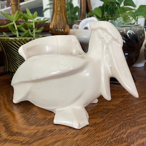 May include: A vintage off-white ceramic pelican figurine. The pelican has a long beak and is perched on a flat base. The figurine is likely a decorative item for a shelf or table.