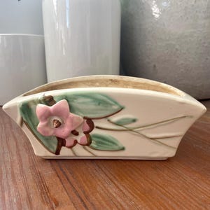 May include: Cream-colored ceramic planter with a floral design. The planter has a rectangular shape with a curved top and is decorated with pink flowers and green leaves. The planter is sitting on a wooden surface.