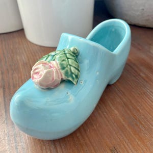 May include: A light blue ceramic clog-shaped planter with a pink and green floral accent. The planter is a decorative item, likely for small plants or flowers. The clog is a traditional Dutch design.