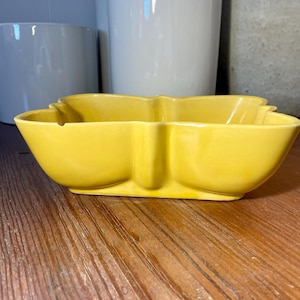 May include: A bright yellow, ceramic, three-sectioned dish with a unique, stylized shape. The dish is sitting on a wooden surface, with white ceramic pots in the background.