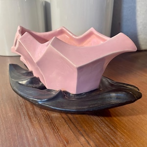 May include: A pink geometric ceramic planter sits on a dark gray, leaf-shaped ceramic base. The planter has a unique, angular design. The background includes white ceramic pots and a wooden surface.