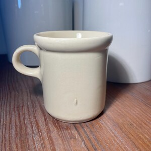 May include: A small, off-white ceramic mug with a rounded rim and a small handle. The mug is cylindrical and sits on a wooden surface. In the background are white ceramic planters.