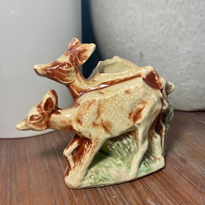 May include: A ceramic figurine of two deer, one atop the other, in a cream and brown color scheme. The base features green painted grass. The figurine has a cracked glaze finish.