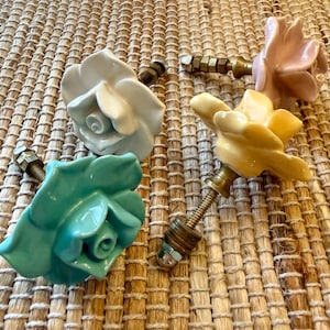 May include: Four ceramic rose-shaped cabinet knobs in white, pink, yellow, and teal. Each knob has a brass-colored threaded stem with a nut for attachment. The knobs are arranged on a woven, light brown surface.