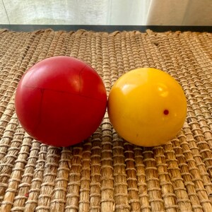 May include: Two spherical objects: a solid red ball and a yellow ball. The red ball has a crack across its surface. The yellow ball has a small red dot. Both balls are on a woven, brown surface.