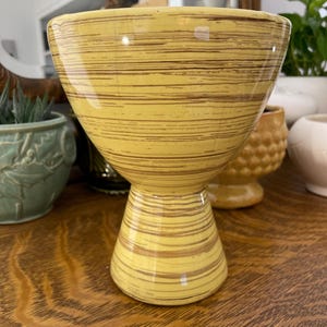 May include: A yellow ceramic planter with a brown horizontal stripe pattern. The planter has a wide, bowl-like top and a pedestal base. The planter is approximately 6 inches tall.