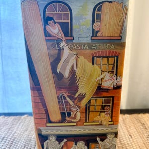 May include: A rectangular metal pasta tin with a detailed illustration of a building facade. The artwork depicts people hanging pasta from windows, with the text "LA PASTA ATTICA" visible. The tin has a decorative roof and base.