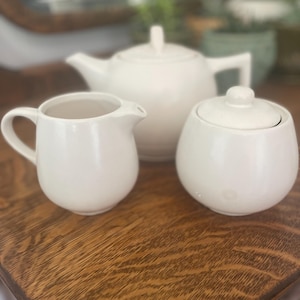 May include: A white ceramic tea set, including a teapot, creamer, and sugar bowl, is arranged on a wooden surface. The teapot has a lid and a handle. The creamer has a handle and a spout. The sugar bowl has a lid.