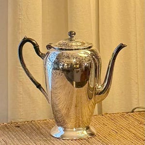 May include: A shiny silver teapot with a curved spout and a black handle. The teapot has a lid with a small knob and sits on a pedestal base. The teapot is reflective, showing the surrounding environment.