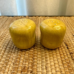 May include: A pair of yellow ceramic salt and pepper shakers. Each shaker has a rounded shape with a glossy finish and several small holes on top. The shakers sit on a woven, textured surface.