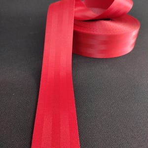 May include: Red webbing with a striped pattern. The webbing is rolled up and the end is laying flat.