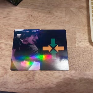 May include: Two rectangular cards with a holographic finish. One card features a photo of a person, the other has a black background with green and orange arrow graphics. The cards are on a wooden surface.