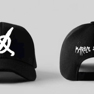 Puede incluir: Gorra de béisbol negra con un gráfico blanco en la parte delantera y las palabras "Marble Hornets" en la parte posterior. El gráfico frontal es un símbolo blanco. La gorra está hecha de tela oscura.