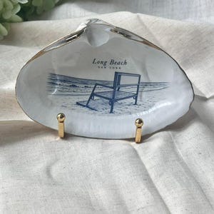 May include: A white seashell trinket dish with a gold-painted rim, displayed on a small gold metal stand. The interior features a blue-toned illustration of a lifeguard chair on a beach with the text Long Beach New York printed above the image.