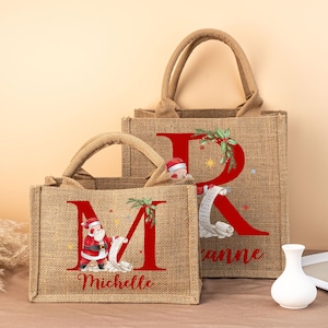 Puede incluir: Dos bolsas de yute con asas. Las bolsas presentan una gran letra roja, un gráfico de Papá Noel y los nombres "Michelle" y "Rianne". Las bolsas están decoradas con elementos temáticos navideños.