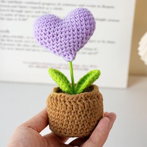 May include: A handcrafted crochet flower in a small pot. The flower features a purple heart-shaped bloom, green leaves, and a green stem. The pot is brown and the piece is made of yarn.