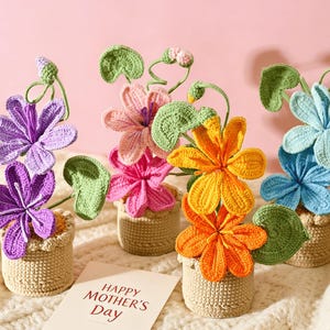 以下が含まれることがあります： 紫、ピンク、オレンジ、青など、さまざまな色の手編みの花が、小さなベージュの手編みの鉢に飾られています。緑の葉と茎が花に添えられています。「HAPPY MOTHER'S Day」と書かれたカードが見えます。