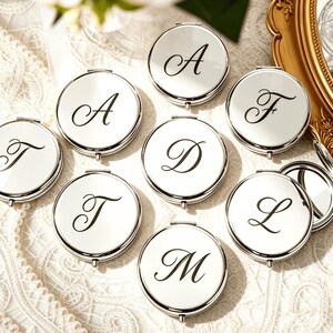 Puede incluir: Una colección de espejos de bolsillo redondos de metal plateado, cada uno con una letra negra en fuente cursiva. Las letras A, D, F, L, M y T son visibles en las superficies pulidas de estos pequeños accesorios personales con bisagras.