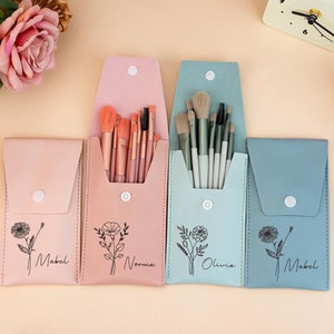 Op de afbeelding: Vier make-up kwastensets in pastelroze, lichtblauwe en blauwe etuis. Elke etui heeft een bloemenontwerp en een naam erop gedrukt. De etuis zijn open en onthullen de kwasten binnenin. Een klok staat op de achtergrond.