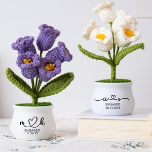 Pode incluir: Dois arranjos de flores de crochê em vasos brancos. Um apresenta flores roxas com centros amarelos, o outro flores brancas com centros amarelos. Os vasos têm datas e iniciais de noivado. As flores têm caules e folhas verdes.