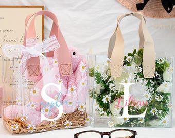 May include: Two clear plastic tote bags with white daisy print and personalized with the letters "S" and "L". The bag on the left contains a pink plush bunny. The bag on the right has a white background with a daisy print.