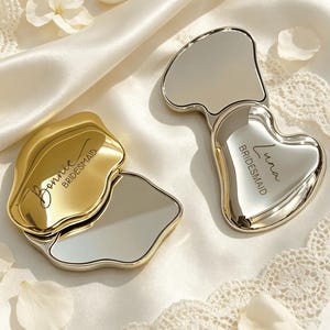 May include: Two compact mirrors with metallic finishes, one gold and one silver. The gold mirror is engraved with the name Bonnie and the word BRIDESMAID. The silver mirror is heart-shaped and engraved with the name Luna and the word BRIDESMAID.