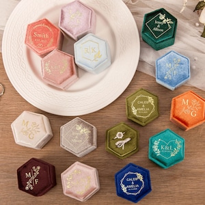 Puede incluir: Una colección de joyeros hexagonales en varios colores, incluyendo rosa, azul, verde y naranja. Cada joyero está personalizado con letras doradas, mostrando iniciales, nombres y fechas. Algunos joyeros están sobre un plato blanco, otros sobre una superficie de madera.