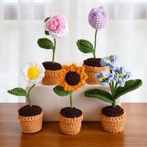 May include: Six crocheted flowers in small terracotta-coloured pots. The flowers include a white daisy with a yellow centre, a pink and white rose, a purple tulip, a yellow sunflower, and blue flowers. Each flower has green leaves and a brown base.