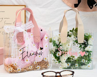 May include: Two clear tote bags with pink handles. One bag contains a pink plush item with daisy accents and the name "Eleanor". The other bag holds white flowers and greenery with the name "Sophia" and a pink heart. A pair of eyeglasses rests in front.