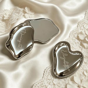 May include: Two silver-toned, irregular-shaped compact mirrors with reflective surfaces. One mirror is engraved with the name Daisy and a small heart, while the other is engraved with the name Elizabeth and a small heart. Both items feature smooth, metallic edges.