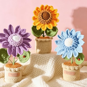 Può includere: Tre fiori all'uncinetto fatti a mano in vasi. I fiori sono di un vibrante viola, giallo e blu, ognuno con foglie verdi e una piccola coccinella. I vasi sono beige con un'etichetta "fatto a mano".